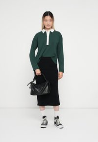 Polo à manches longues vert avec col blanc, jupe noire ajustée, sac à main noir avec motif texturé, chaussettes blanches et baskets noires.