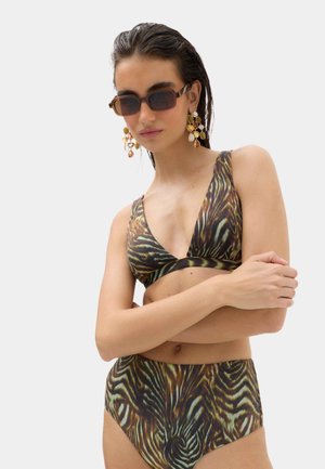 Kvinde med vådt hår iført mønstret bikini, store hængende øreringe og solbriller, stående med armene over kors foran en ensfarvet baggrund.