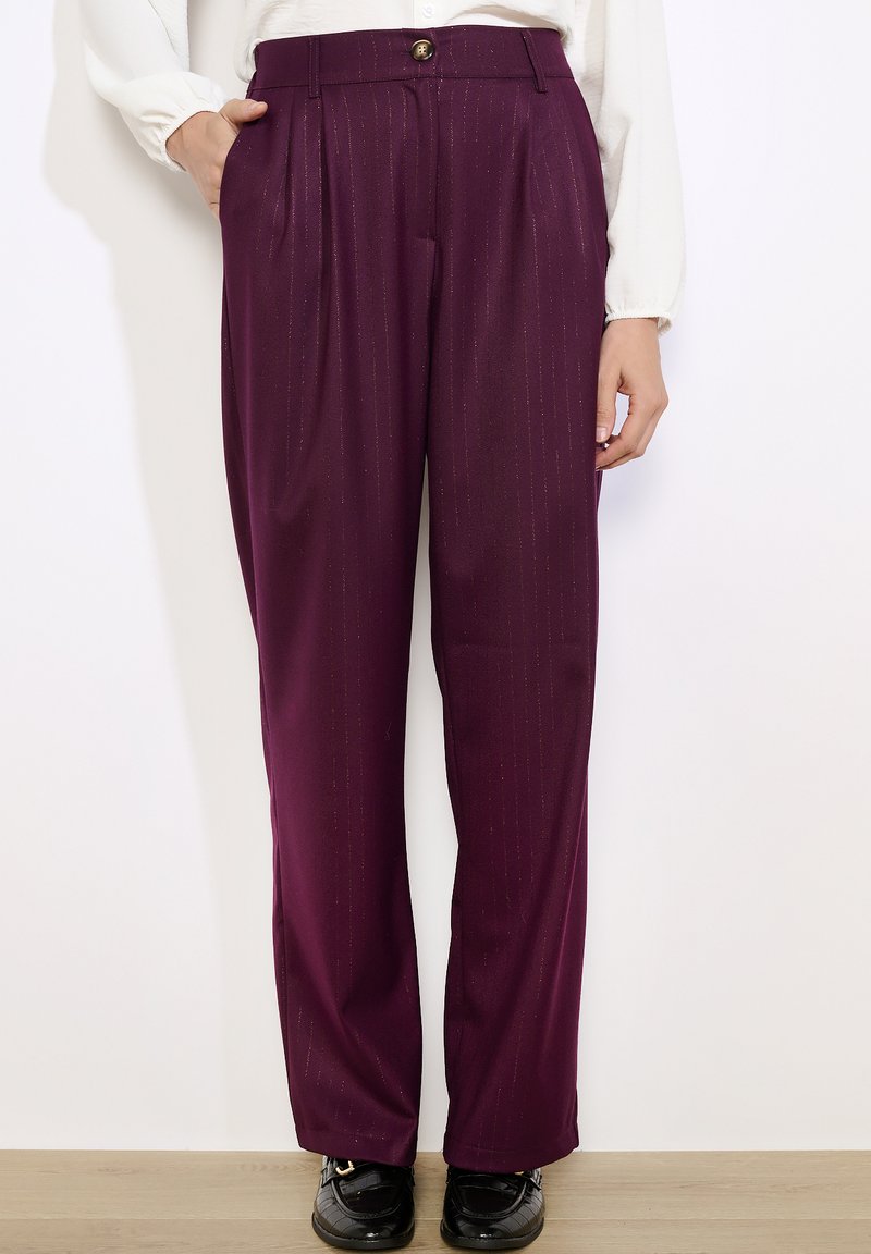 Naine kannab kõrge vöökohaga, sirge lõikega, tumepurpurseid pükse, millel on märkamatu vertikaalne triip, koos mustade lohavede ja valge pika varrukaga t-särgiga.