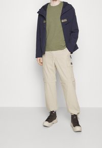 Marine letvægt jakke med hætte, grøn t-shirt, creme cargo bukser og mørkebrune højtop sneakers, med lommer og justerbare manchetter.