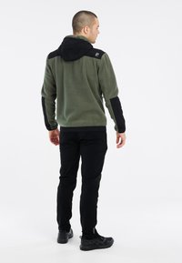Fleece-Hoodie in Olivgrün mit schwarzen Akzenten und Kapuze. Verfügt über eine glatte Textur und einen entspannten Schnitt, kombiniert mit schwarzen Hosen und Schuhen.