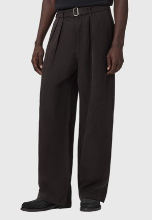 Mann trägt lockere schwarze Hose mit Gürtel und schwarzen Schuhen, steht vor einem einfarbigen hellen Hintergrund.