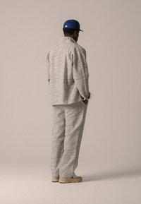 Veste texturée gris clair et pantalon assorti, avec un col haut et une coupe décontractée. Des chaussures beiges et une casquette de baseball bleue complètent la tenue.