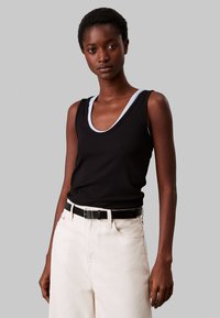 Top in tessuto a coste nero con scollatura profonda, rifinito con un bordo azzurro chiaro, abbinato a jeans a vita alta color crema e cintura nera.