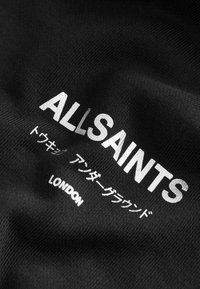 Crna tkanina s teksturiranom površinom sadrži tekst "ALLSAINTS" u bijeloj boji, uz japanski natpis i "LONDON" ispod u manjem bijelom fontu.