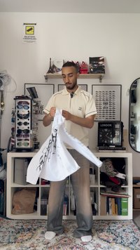 Fehér, hosszú ujjú ing fekete faág mintával, egy személy kezében. Világosszürke nadrág és fehér zokni látható. A háttérben napszemüveg található.