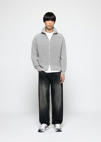 Ung mann med rett, svart hår som har på seg en grå zip-up genser, hvit t-skjorte, svarte, falmede bukser med vidde og hvite sneakers som står.