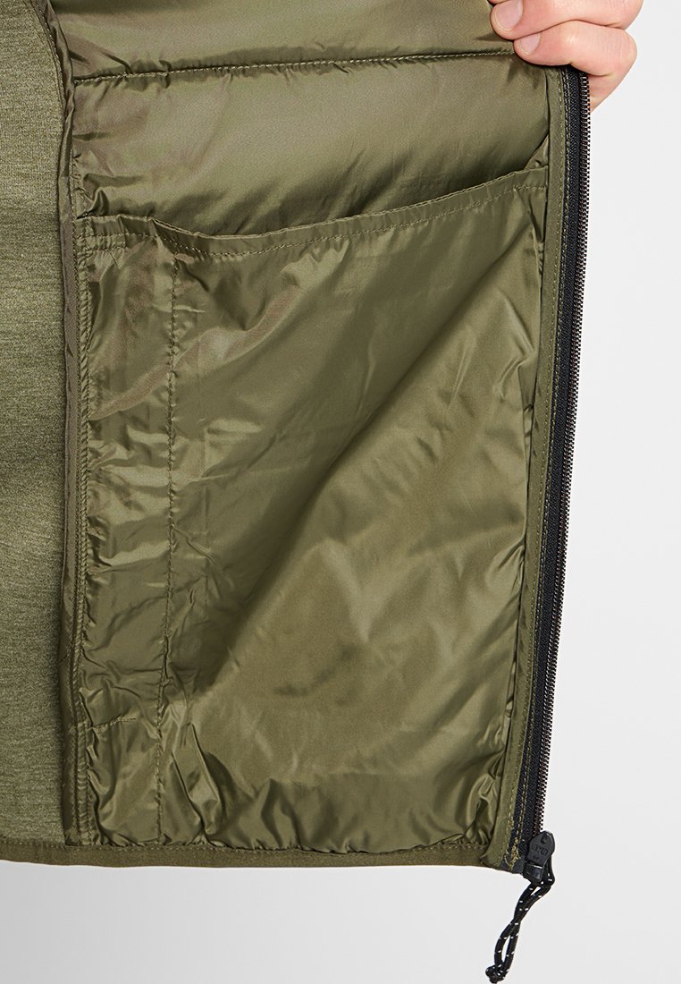 Dłoń trzyma otwartą oliwkowozieloną kurtkę, ukazując gładką podszewkę z przeszywaną kieszenią oraz czarnym suwakiem z zaczepem.