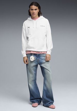 Młody mężczyzna stojący, ubrany w białą bluzę z kapturem Puma Porsche, różową koszulę, luźne, wyblakłe niebieskie jeansy z małą białą torbą przypiętą i różowe sneakersy.