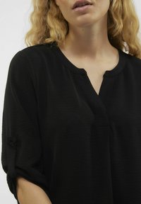 Blusa nera con una texture morbida, scollatura a V e maniche arrotolate con dettagli a bottone. Design semplice, adatta per un abbigliamento casual.