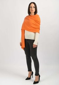 Orange uld sjal lagt over en hvid sweater, parret med sorte stramme jeans og sorte højhælede sko. Modellen står foran en ensfarvet baggrund.