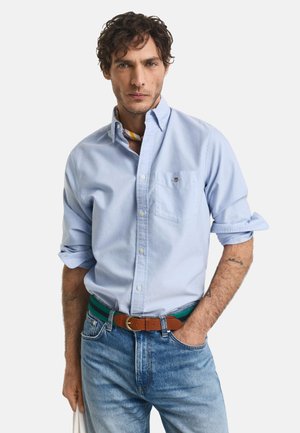 Light blue button-down shirt with rolled sleeves, pocket detail, and a brown belt with green accents, paired with light blue denim jeans.
