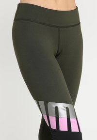 Olivově zelené legíny s vysokým pasem, kontrastními růžovými a stříbrnými grafickými akcenty blízko lemu, vyrobené z elastické, hladké tkaniny.