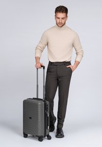 Hombre en un cuello de tortuga beige y pantalones oscuros, de pie con una mano en el bolsillo y la otra sosteniendo la asa de una maleta gris con ruedas.