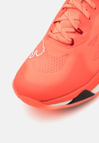 Scarpa sportiva arancione brillante con una tomaia texturizzata, lacci sottili e una suola in gomma con un dettaglio nero. Dettaglio del logo ben visibile in bianco.