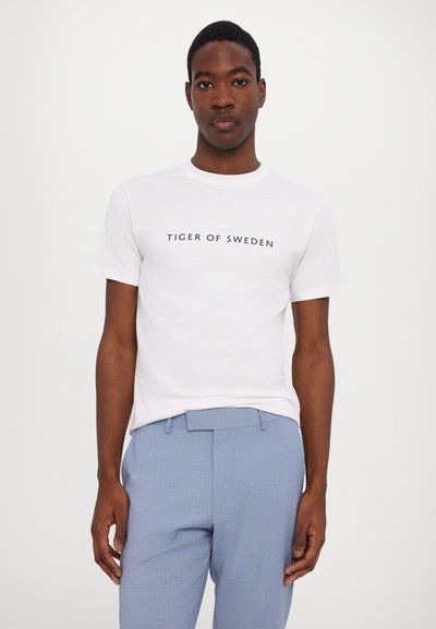 Weißes Baumwoll-T-Shirt mit Rundhalsausschnitt, bedruckt mit "TIGER OF SWEDEN" in dunkler Schrift. Kombiniert mit hellblauen strukturierten Hosen.
