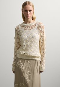 Jersey de color crema, tejido, con un patrón de encaje en forma de diamante y un cuello redondo acanalado. Combinado con una falda beige que presenta diseños de hojas bordadas.