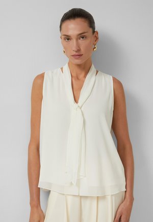 Vrouw met donker haar naar achteren gebonden, met gouden creooloorbellen en een mouwloze witte blouse met een knoopdetail aan de voorkant tegen een grijze achtergrond.