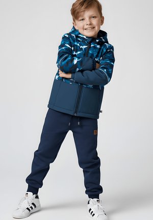 Uśmiechnięty chłopiec stoi z założonymi rękami, ubrany w niebieską kurtkę moro, granatowe dresy i białe trampki na jednolitym szarym tle.