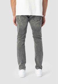 Persona con jeans grises, zapatillas blancas y camiseta blanca, de pie con las manos relajadas a los lados, vista desde atrás.