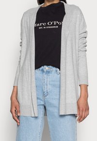 Světle šedý kardigan vyrobený z měkké tkaniny, otevřený vpředu, dlouhé rukávy, žebrované manžety, nošený přes černé tričko a světle modré džíny.