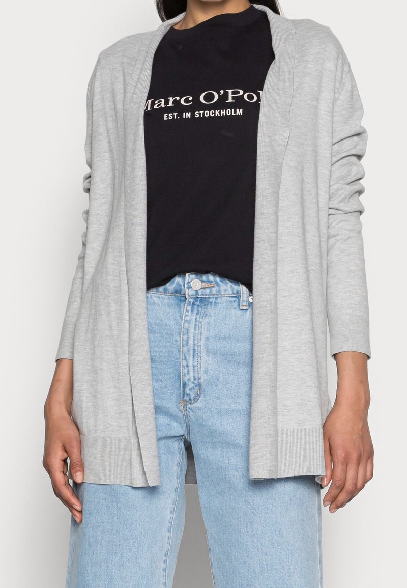 Světle šedý kardigan vyrobený z měkké tkaniny, otevřený vpředu, dlouhé rukávy, žebrované manžety, nošený přes černé tričko a světle modré džíny.