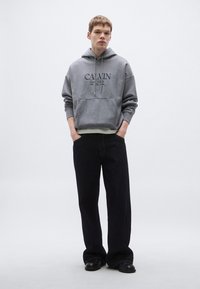 Szürke kapucnis pulóver elülső zsebbel, fekete logóval. Laza fazonú sötét farmerral és fekete cipővel párosítva.