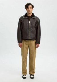 Brun läderjacka med fuskpälskrag, dragkedjestängning och långa ärmar; kombinerad med beige byxor och svartvita skor.