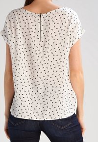 Blusa a maniche corte color crema con un motivo a foglie nere, caratterizzata da un collo rotondo e da una cerniera metallica visibile sul retro.