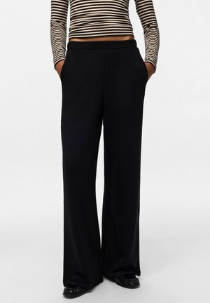 Personne portant un pantalon noir à jambes larges avec les mains dans les poches et un top court à manches longues rayé, debout devant un fond uni.
