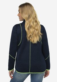 Giacca in pile blu navy con cuciture verdi a contrasto, design allungato e orlo arrotondato, caratterizzata da maniche lunghe e dettagli laterali.