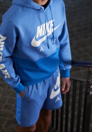 Person trägt einen blauen Nike Air Hoodie und passende Shorts, steht neben einem Metallgeländer, die Hände teilweise in den Taschen.