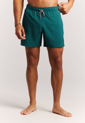 Homme portant un short de bain vert sarcelle avec un cordon noir et blanc, debout pieds nus devant un fond clair uni.