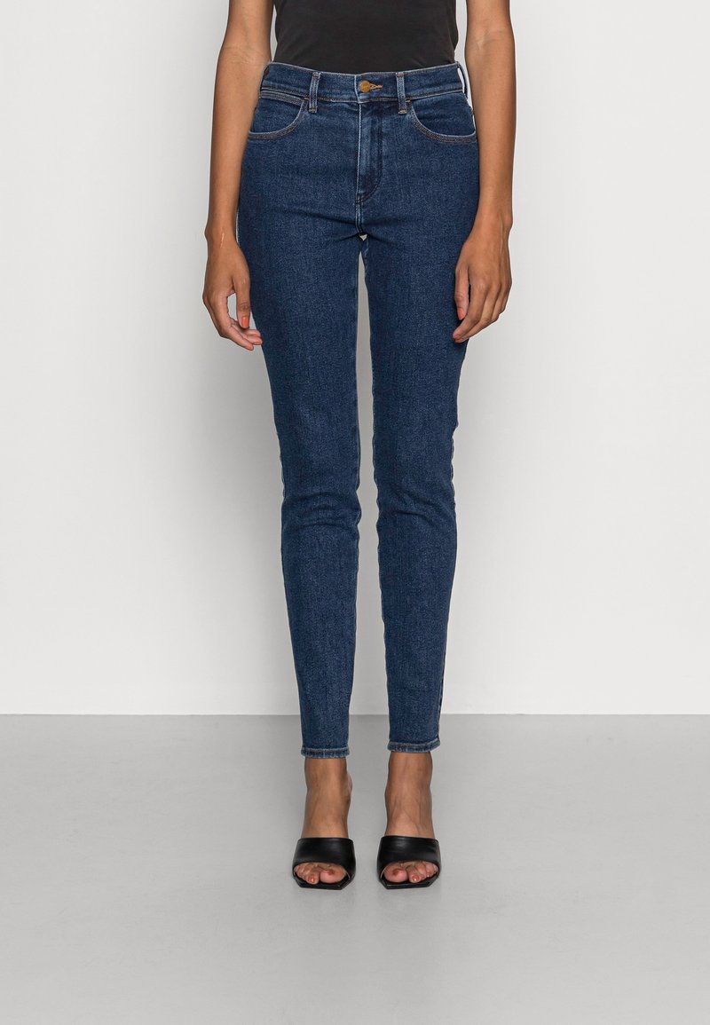 Mujer vestida con vaqueros ajustados de color azul oscuro y tacones abiertos negros, de pie contra una pared blanca lisa y un suelo gris claro.
