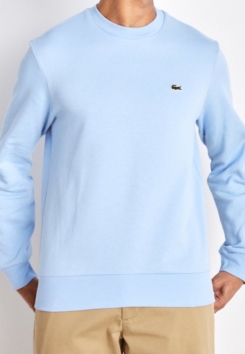 Sweatshirt bleu clair avec un col rond, des poignets et un ourlet côtelés. Présente un petit logo vert sur la poitrine. Fabriqué en tissu doux.