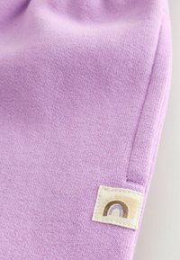 Lilla strikket stof med en blød tekstur. Har et firkantet lappet stykke med et regnbuedesign i bløde farver. Afrundede kanter.