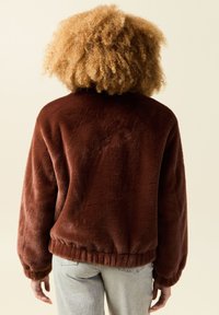 Brązowa kurtka z polaru o luźnym kroju, z prążkowanym wykończeniem i miękką teksturą, pokazana z tyłu, zestawiona z jasnymi dżinsami.