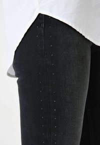 Jeans en denim noir avec une coupe slim, ornés de rangées verticales de petits embellissements sombres le long de la couture extérieure. Le bas d'une chemise blanche dépasse.