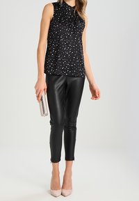 Svart ärmlös topp med vitt stjärnmönster, åtföljd av tajta svarta faux läderleggings och beige högklackade skor, hållandes en silverfärgad kuvertt.