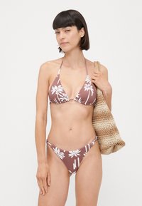 Bikini marrone con motivo di foglie di palma bianche; top triangolare con spalline da annodare, slip a vita bassa coordinati. Modella che tiene una borsa di paglia intrecciata.
