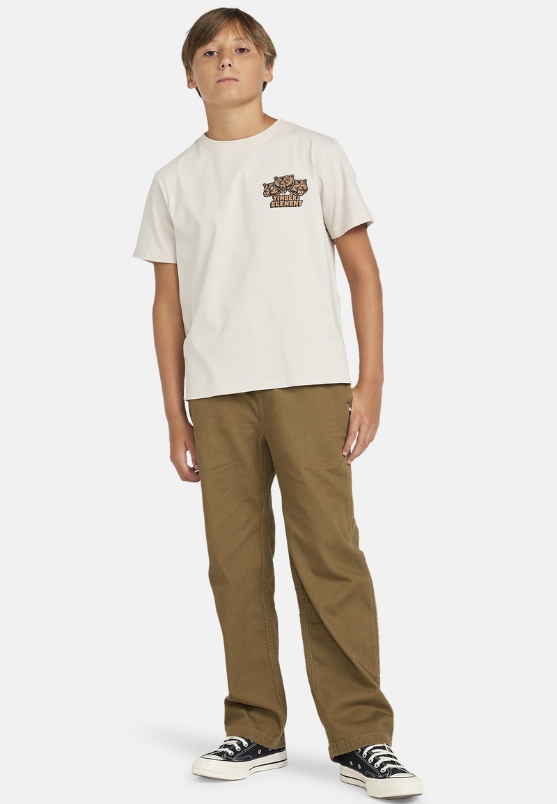 Kermakuvilla t-paita vasemmassa rinnassa olevalla graafisella printillä, yhdistettynä khaki-värisiin puuvillahousuihin ja mustiin kankaasta tehtyihin tennareihin.