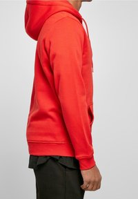 Vista lateral de una persona que lleva una sudadera con capucha roja brillante y pantalones negros, contra un fondo gris claro liso.