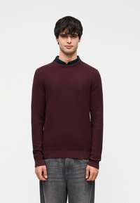 Pull en tricot bordeaux à col rond, présentant un tissu texturé et des poignets côtelés, porté sur une chemise à col foncé, associé à un jean gris.