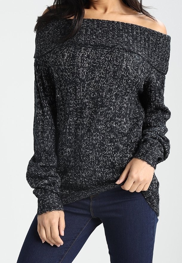 Femme portant un pull en maille noir et blanc chiné, épaules dénudées, et un jean bleu foncé, une main reposant sur sa cuisse.