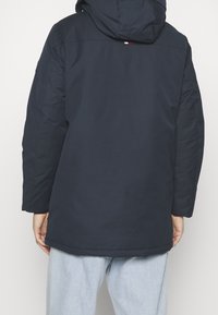 Laivastonsininen toppatakki, jossa huppu, selässä logoyksityiskohta ja joustavat hihansuut, yhdistetty vaaleansinisiin farkkuihin.