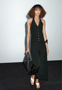 Schwarzes trägerloses Top mit tiefem V-Ausschnitt und Knopfdetails, kombiniert mit weiten, zerknitterten schwarzen Hosen und weißen Sandalen. Geflochtener schwarzer Beutel.