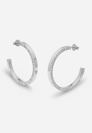 Paire de boucles d’oreilles créoles polies, de ton argenté, ornées de pierres transparentes le long des courbes intérieures, sur un fond clair uni.