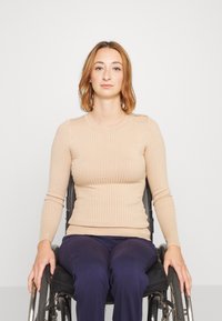 Haut à manches longues beige à côtes associé à un pantalon bleu marine. Modèle assis dans un fauteuil roulant, avec les cheveux raides et un fond neutre.