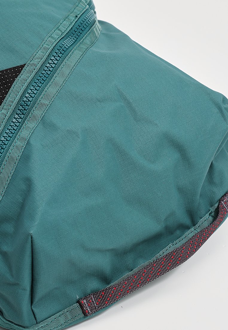 Türkisfarbener Stoffrucksack mit strukturierten Oberflächen, einem Reißverschlussfach und kontrastierenden dunkelgrauen sowie gemusterten Akzenten entlang der Ränder.