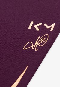 Violetinė medžiaga su tekstūruota paviršiumi. Oranžiniai atspaudai apima stilizuotus rašmenis ir parašą, su įstrižiniu dizaino elementu.